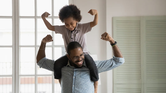 image of a Black father with his son sitting on his shoulders and both making the strong arms pose